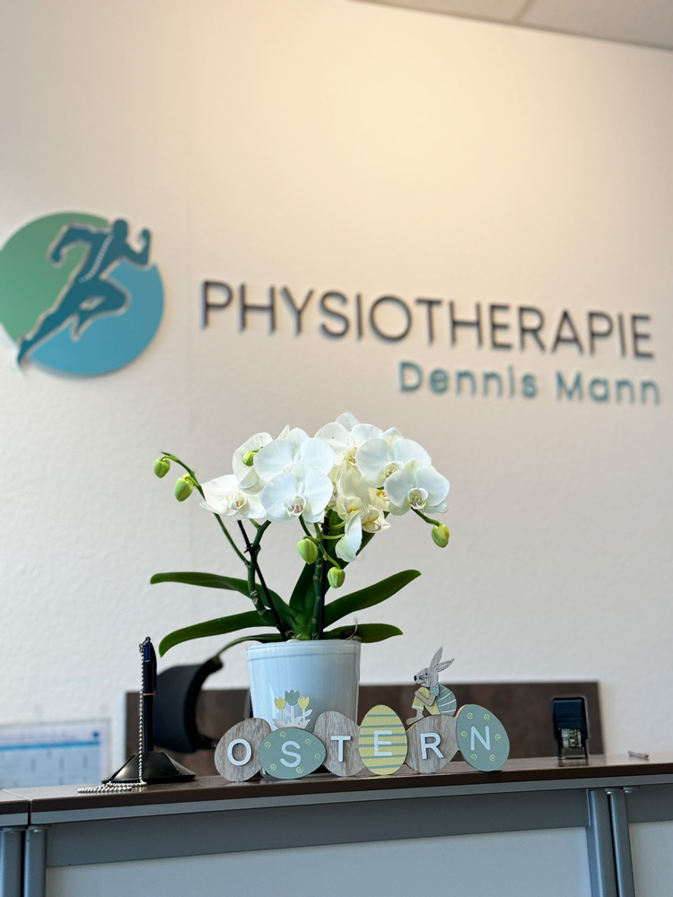 Freundliche Osterdekoration mit Orchidee am Empfang der Physiotherapie Dennis Mann in Mannheim, mit Praxislogo im Hintergrund.